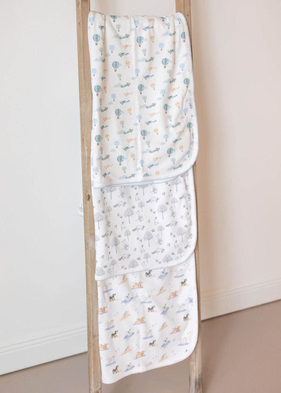 Two baby blankets with a floral pattern draped over a wooden ladder against a white wall.