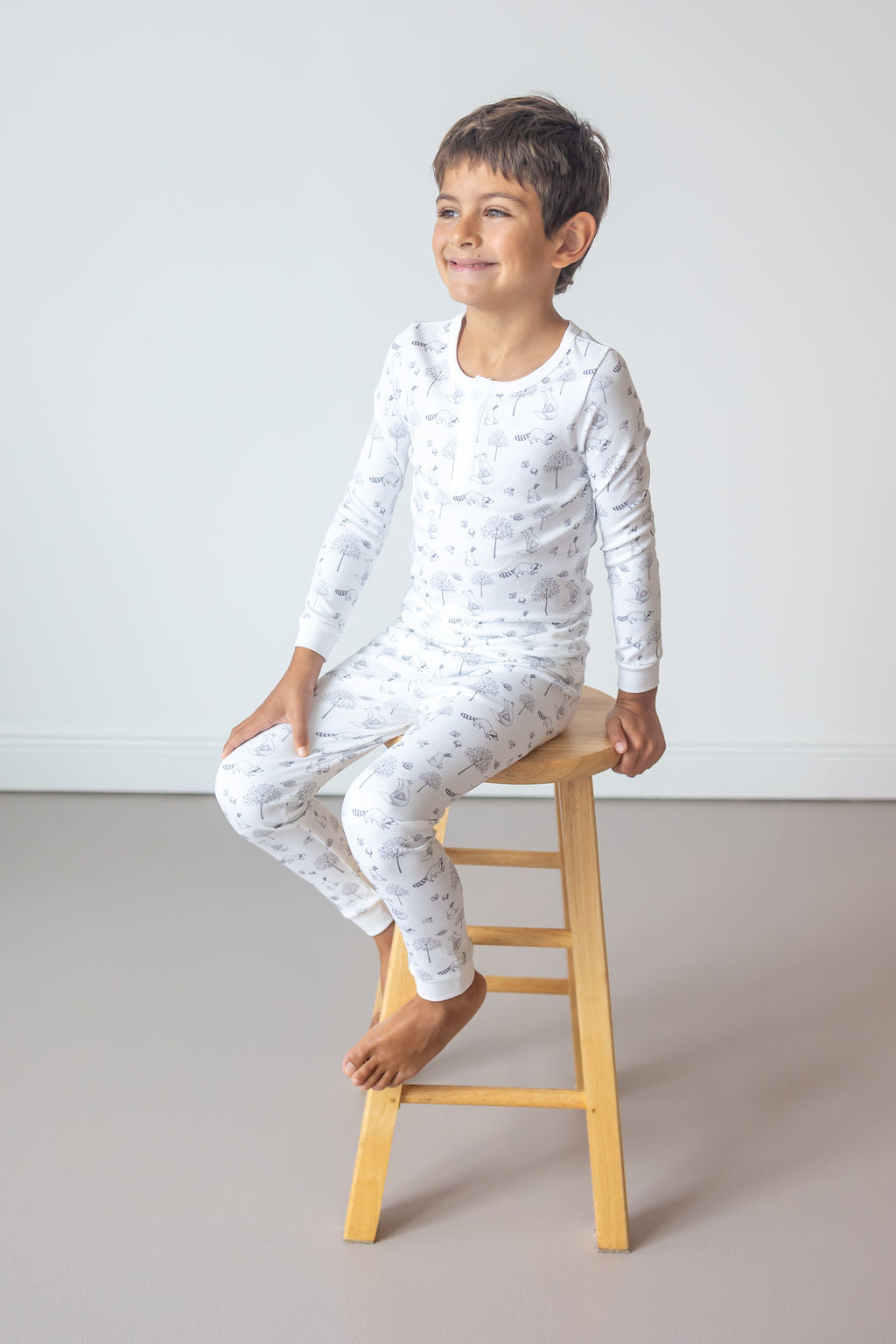 Child wearing a white long-sleeve shirt and pants with small patterns, sitting on a wooden stool against a plain background.