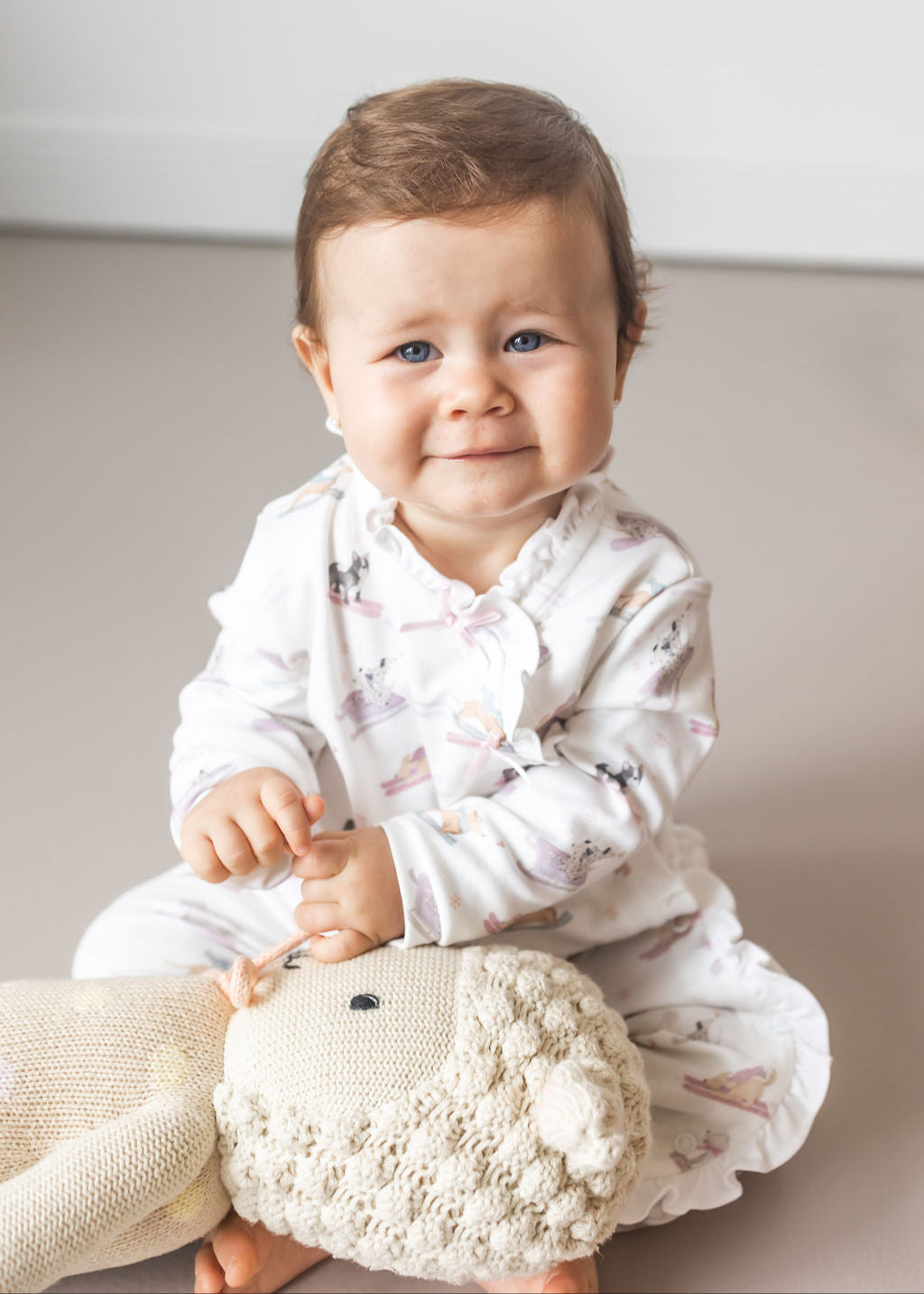 Baby sitting on a cushion holding a plush toy with a plain background