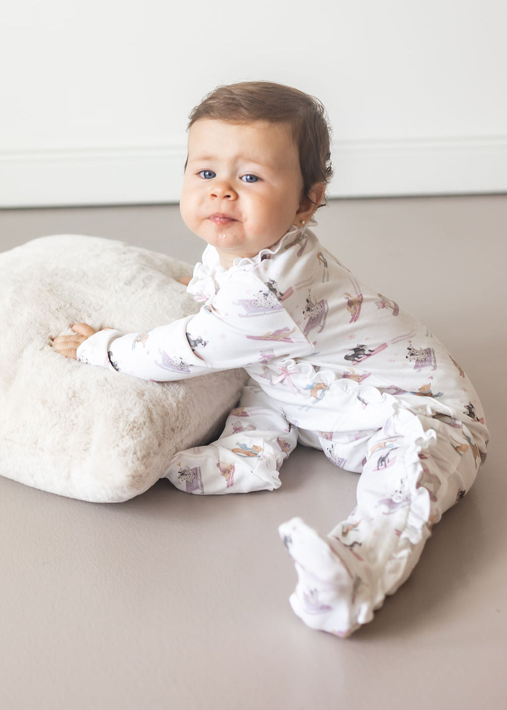 Baby in a white onesie with animal prints sitting on a light-colored surface.