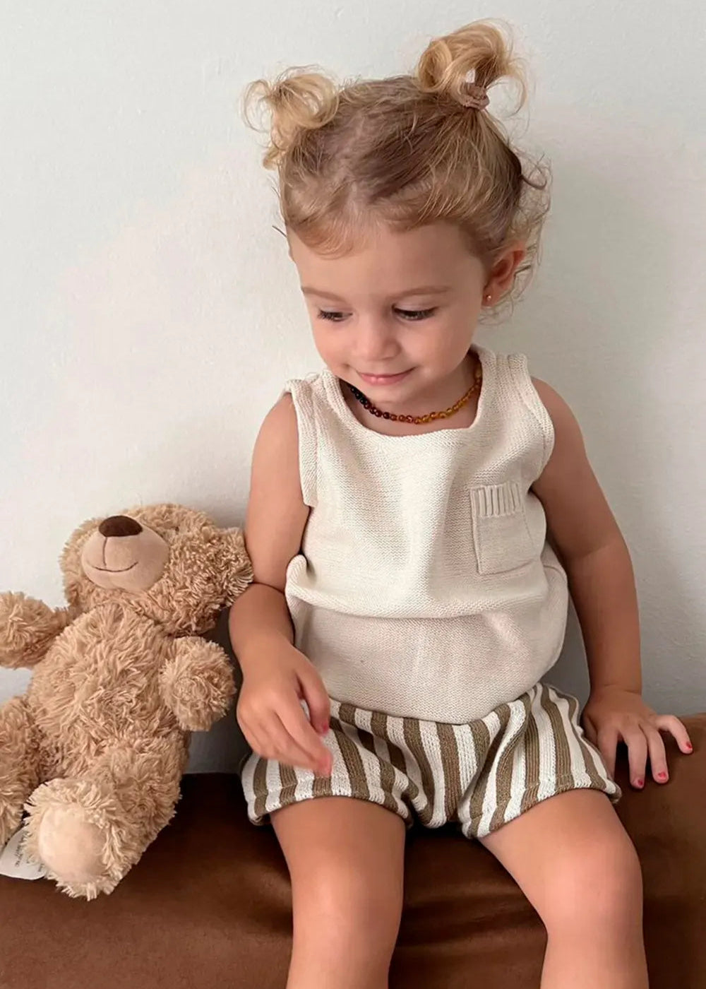 Child sitting with a teddy bear, wearing a white sleeveless top and striped shorts.