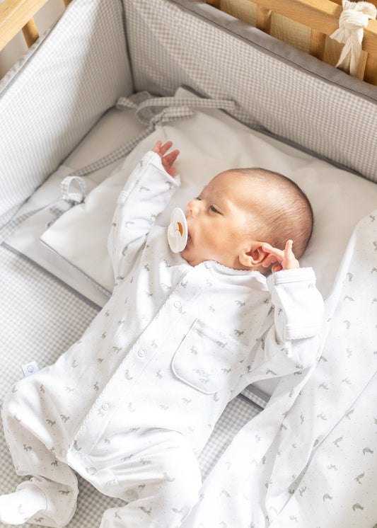 Baby in a crib wearing a white onesie with small patterns, surrounded by soft bedding.