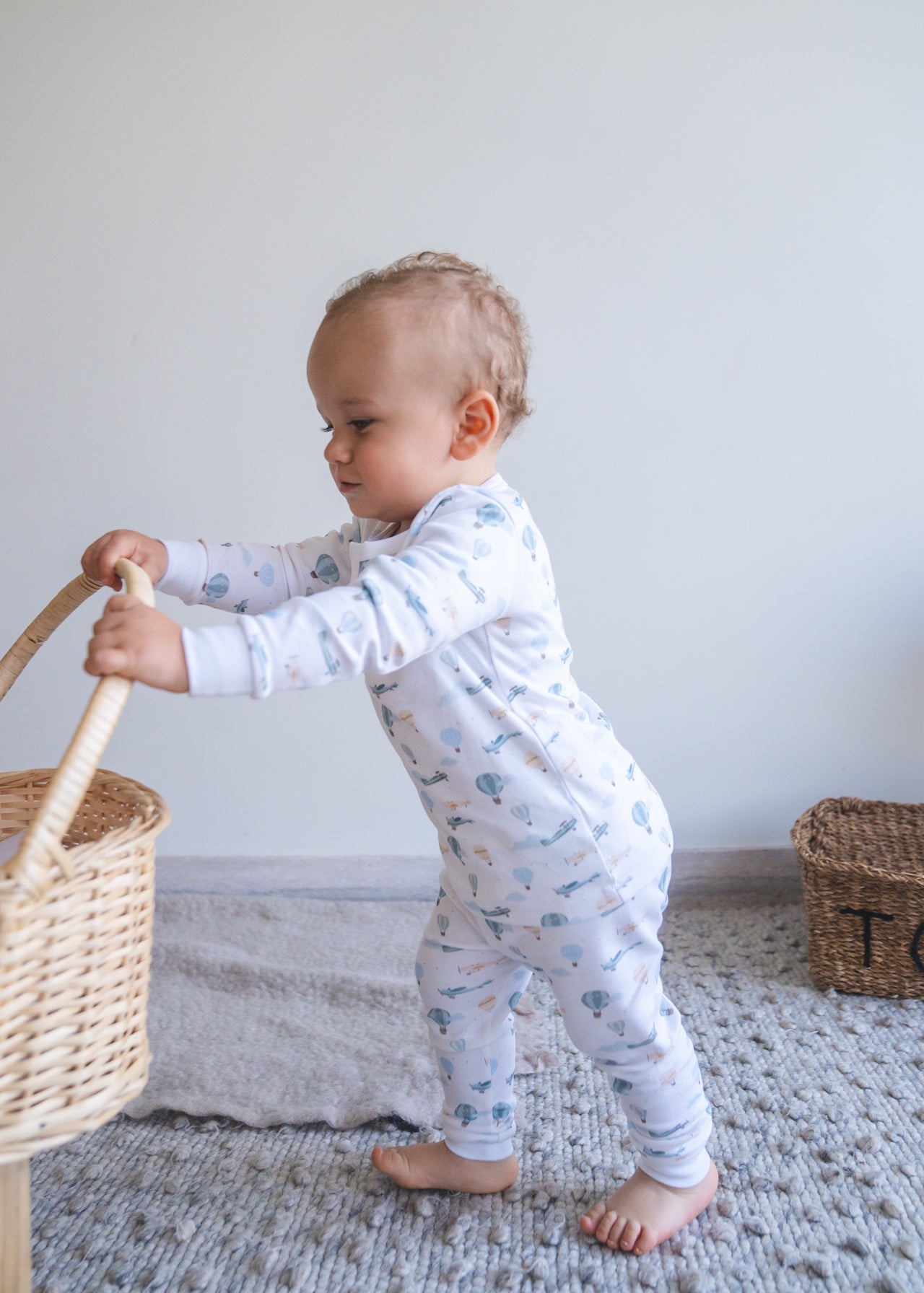 Baby in a light blue onesie with animal prints standing next to a woven basket on a textured rug.