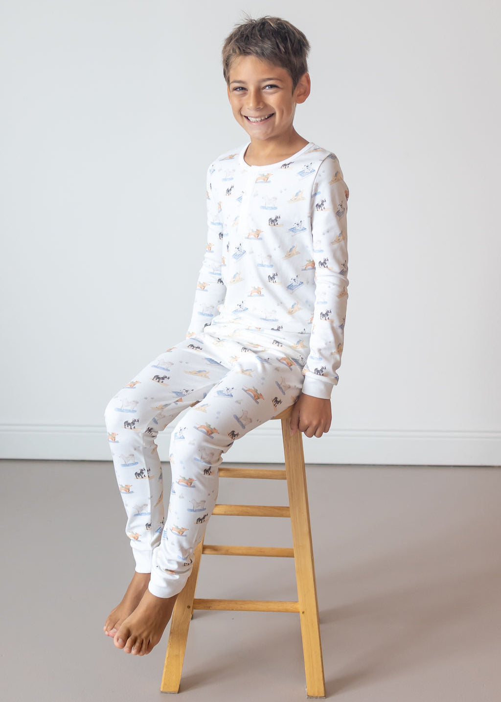 Child wearing a white pajama set with a pattern, sitting on a wooden stool against a plain background.