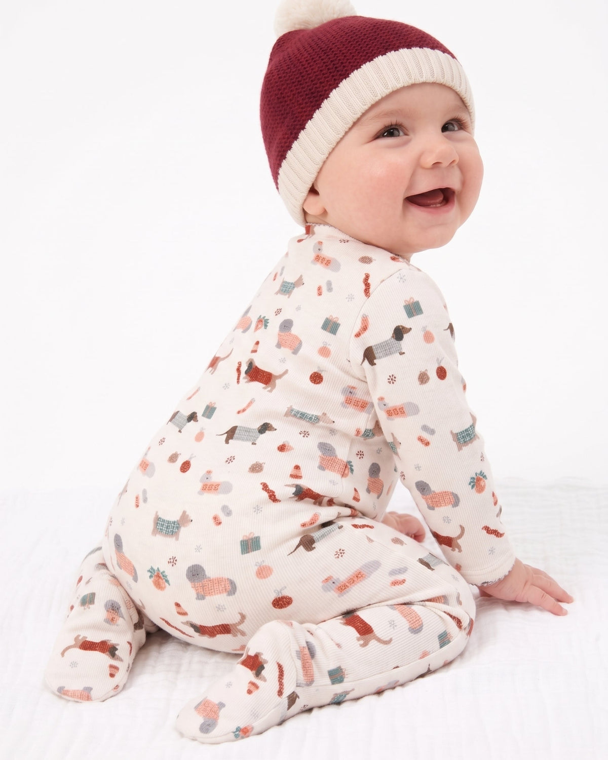 A baby wearing a cream footed sleeper with a multicolor holiday print and a maroon and cream knit pom-pom hat, sitting against a white background.