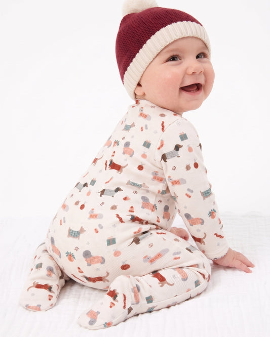 A baby wearing a cream footed sleeper with a multicolor holiday print and a maroon and cream knit pom-pom hat, sitting against a white background.