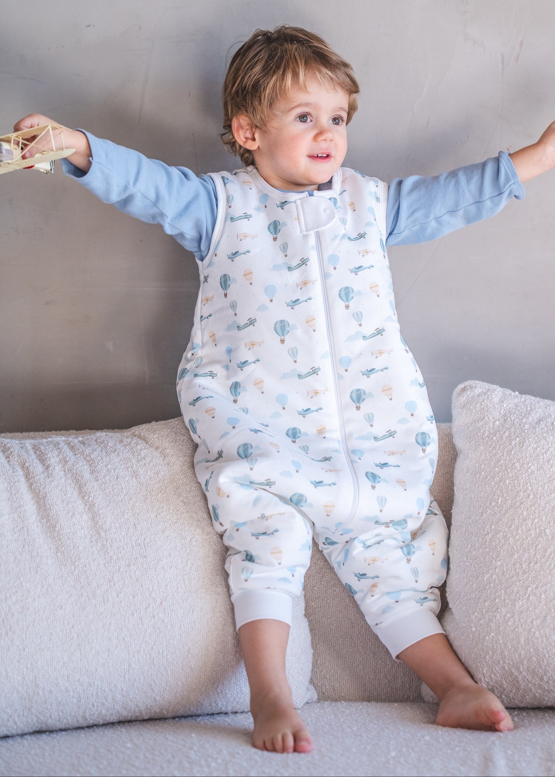 Child in a white onesie with blue sleeves sitting on a couch holding toys.