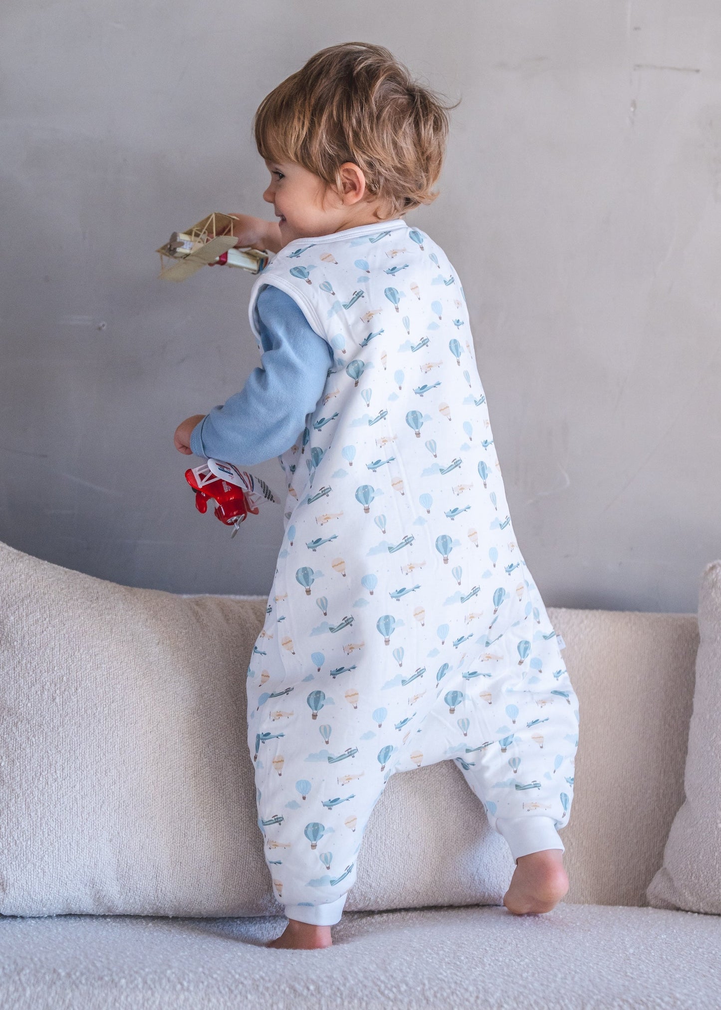 Child in a patterned onesie standing on a couch against a plain wall.