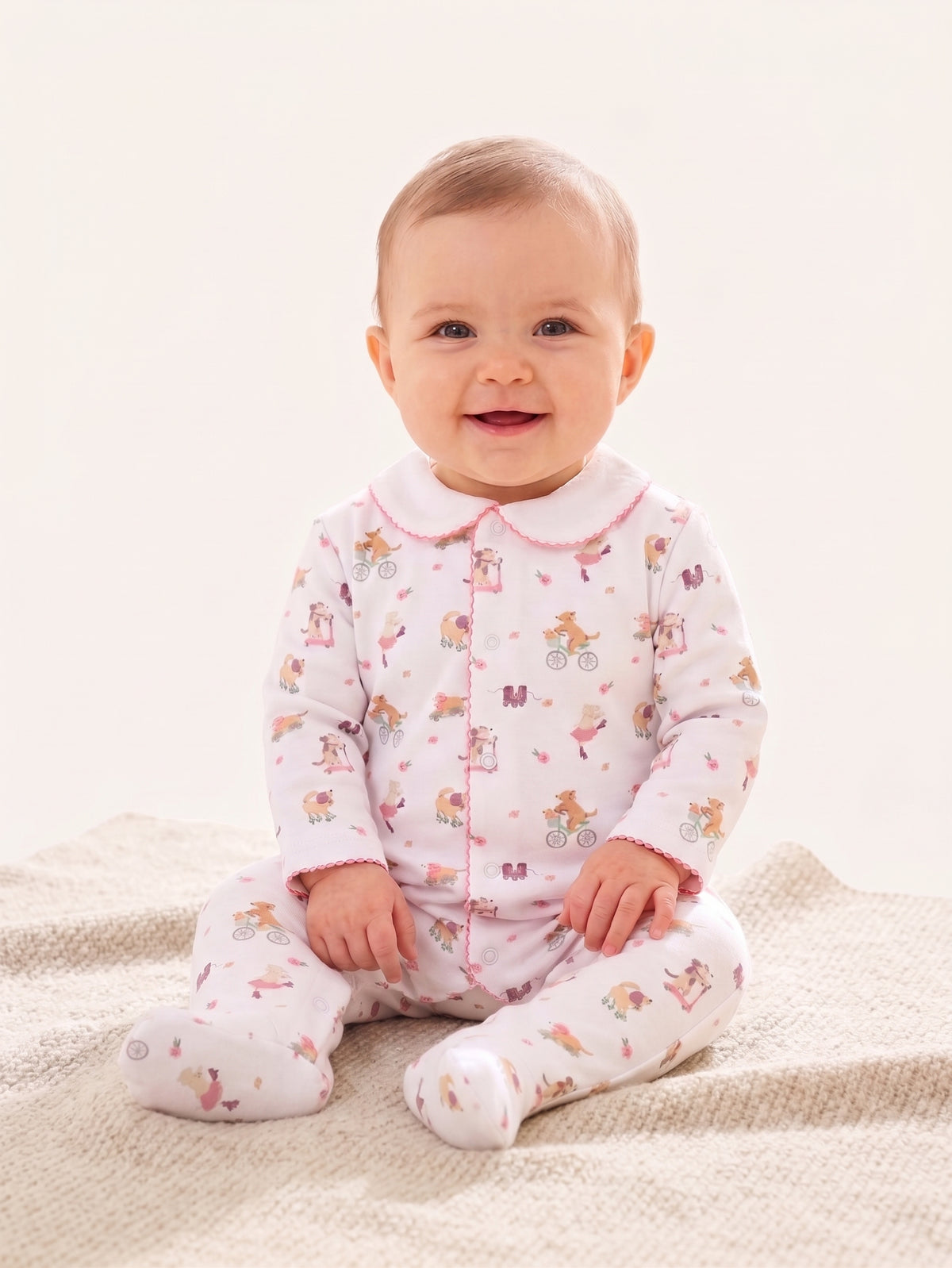 A baby sitting on a textured cream blanket wearing a light pink footed sleeper with a white collar and a colorful cartoon bicycle print.