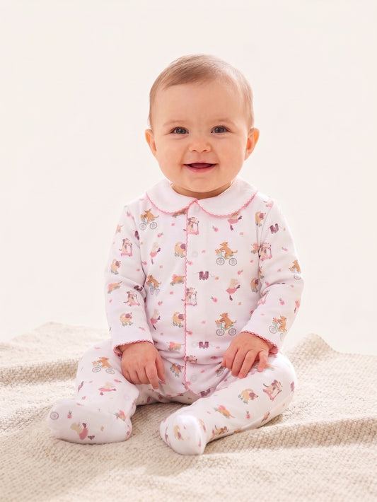 A baby sitting on a textured cream blanket wearing a light pink footed sleeper with a white collar and a colorful cartoon bicycle print.