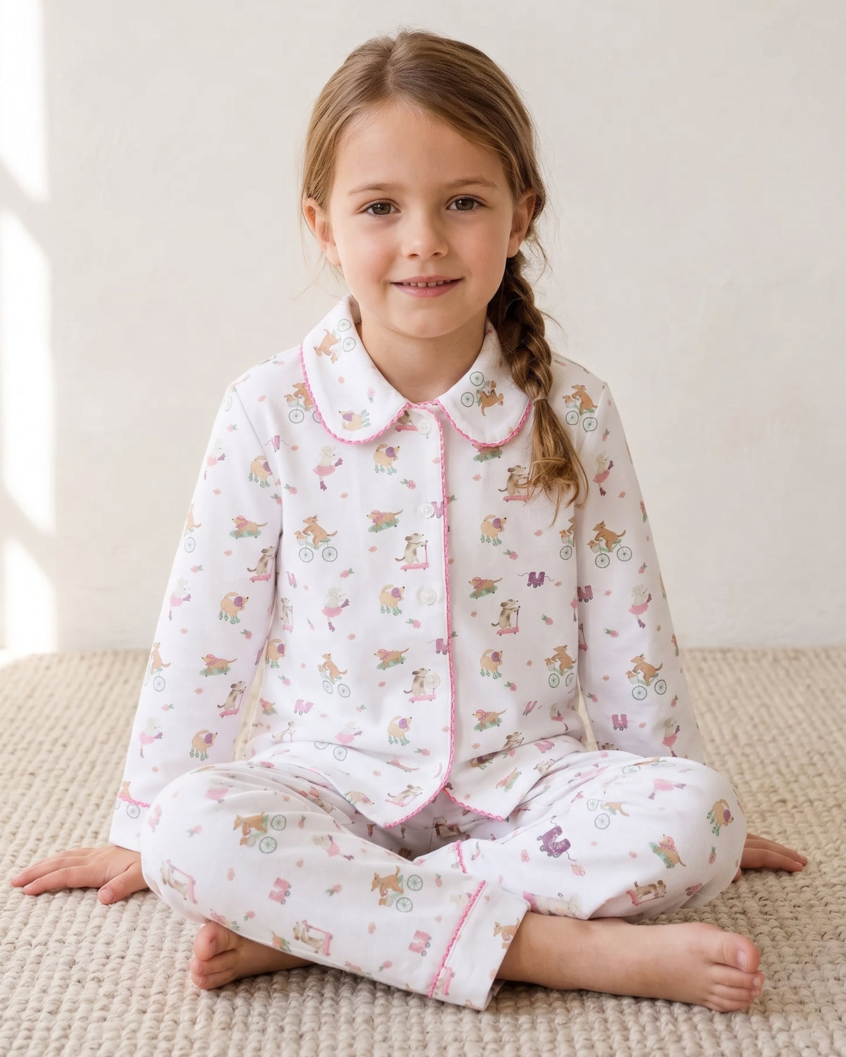 A young girl with a braided ponytail sits cross-legged on a textured beige rug, wearing a white long-sleeve pajama set with pink trim and a colorful cartoon animal print.